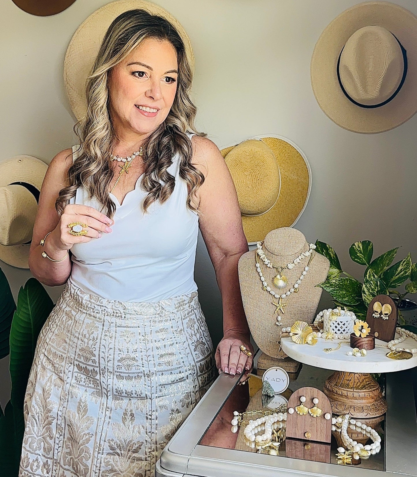 Woman standing next to a display of jewelry and decorative items with hats on the wall behind her.