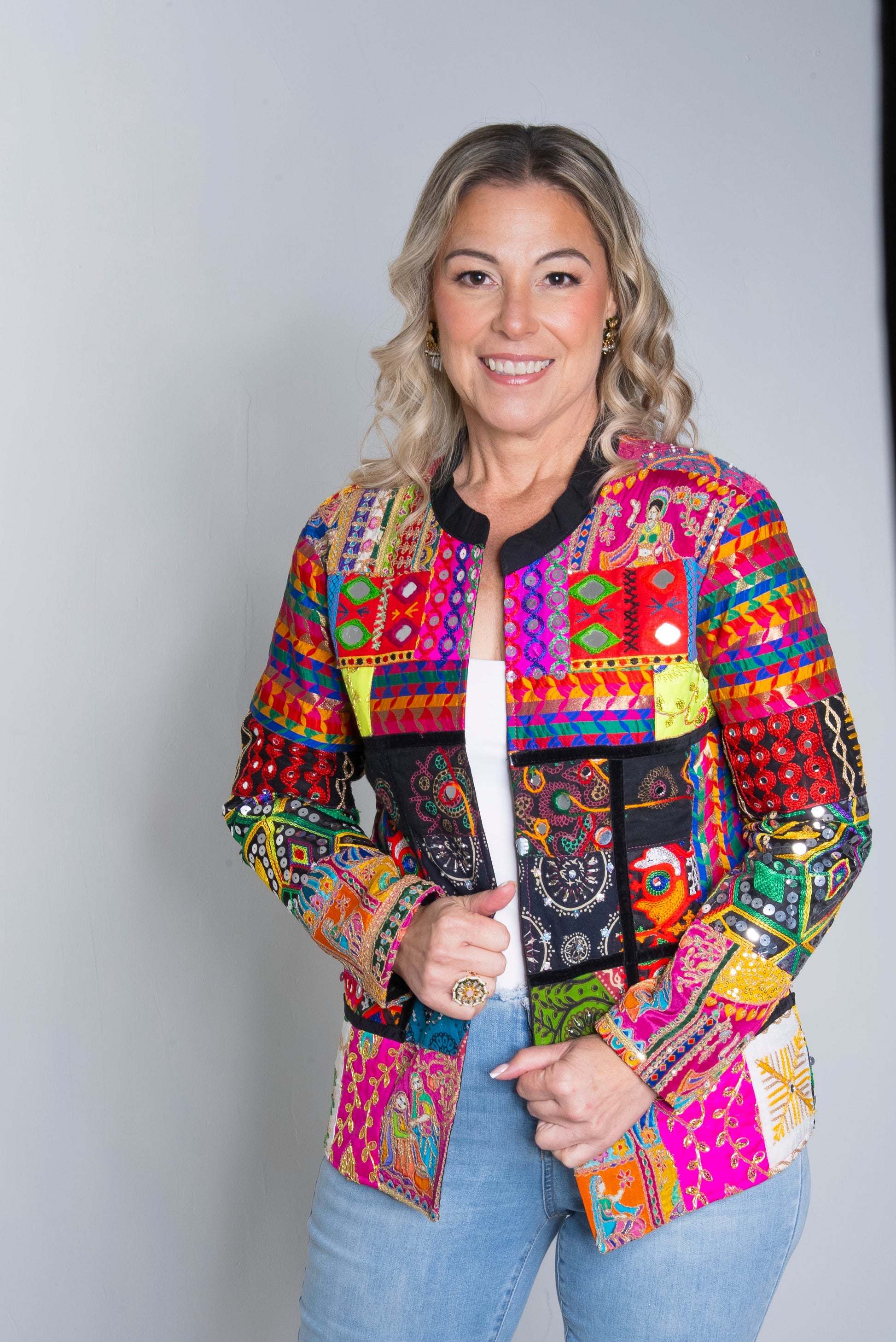 A woman modeling a vibrant, multicolored patchwork jacket with Mexican-style embroidery and geometric patterns, paired with light blue jeans, against a light gray studio background.