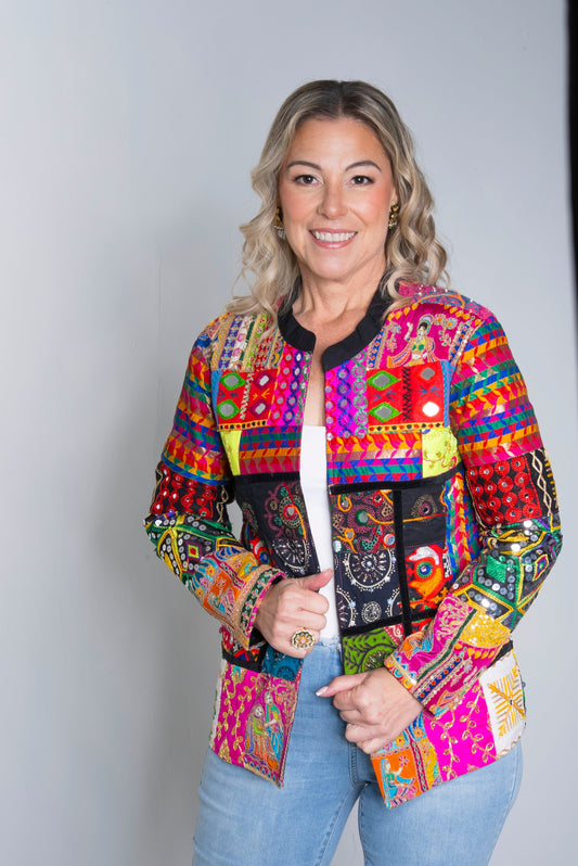 A woman modeling a vibrant, multicolored patchwork jacket with Mexican-style embroidery and geometric patterns, paired with light blue jeans, against a light gray studio background.