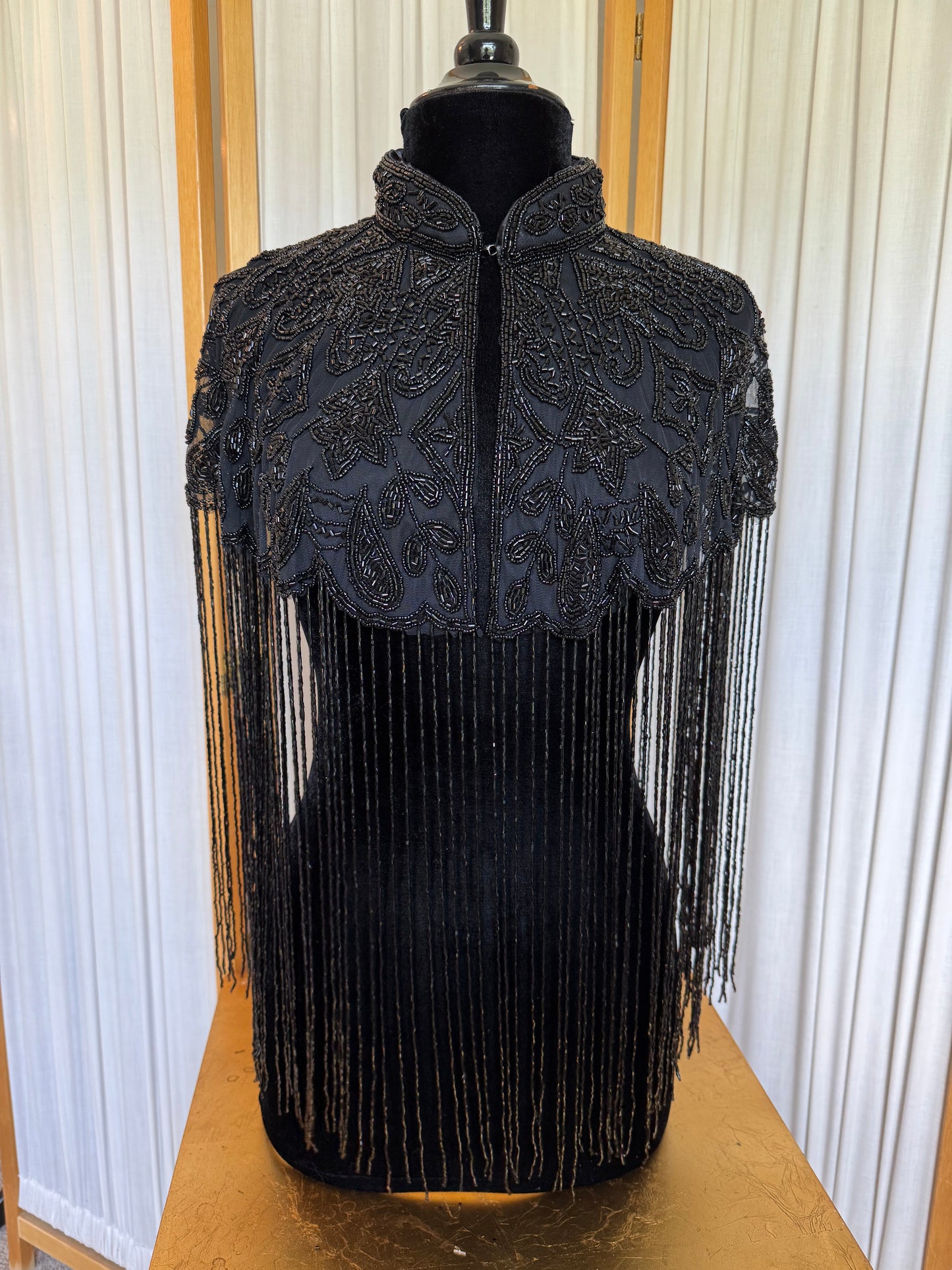 Black beaded and fringed garment on a mannequin against a white curtain background