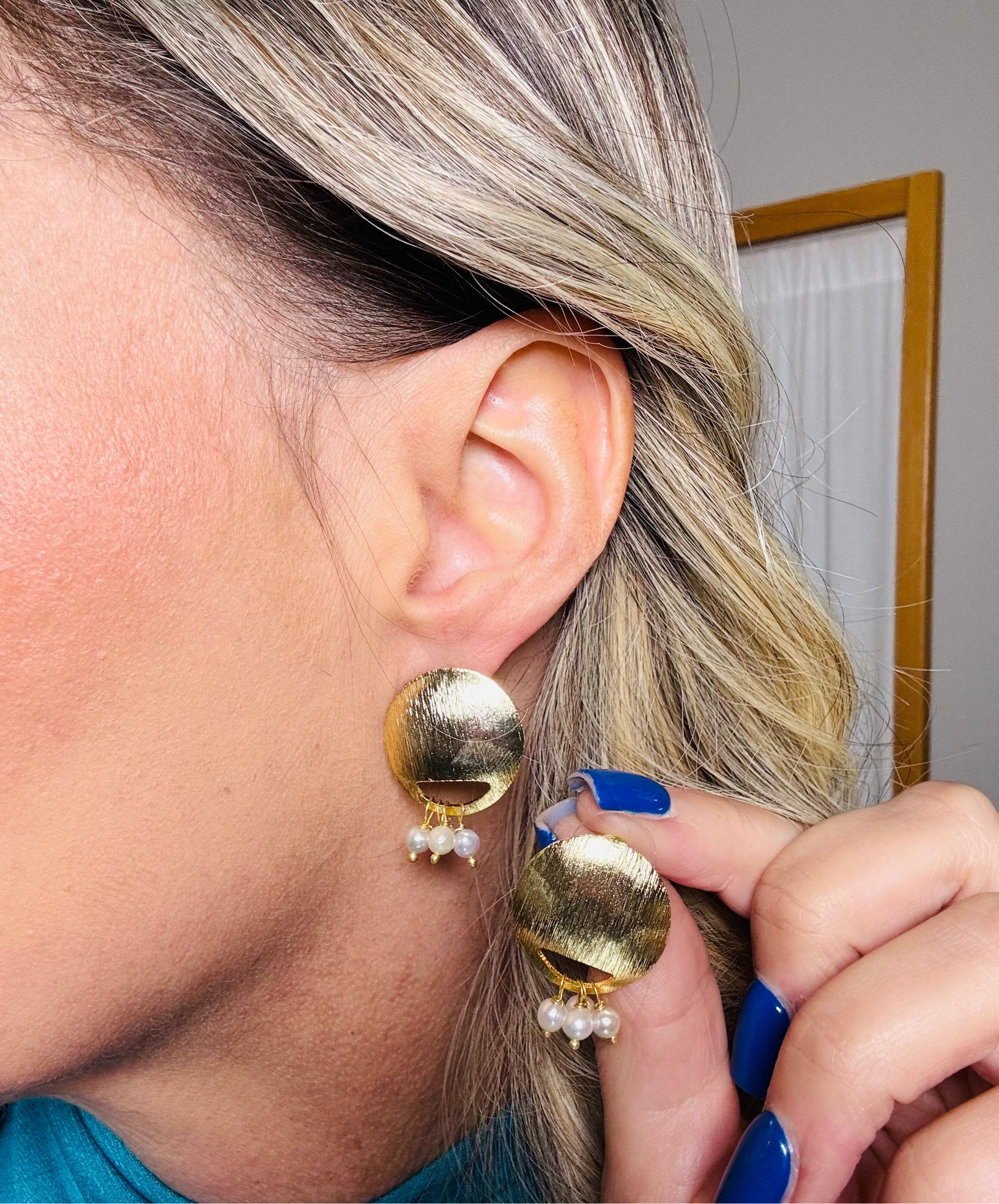 Close-up of a person wearing gold earrings with pearl-like stones, holding a mirror.