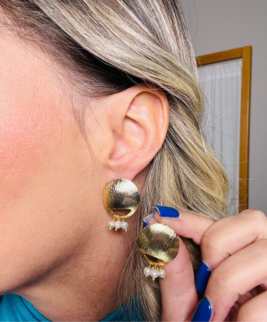 Close-up of a person wearing gold earrings with pearl-like stones, holding a mirror.